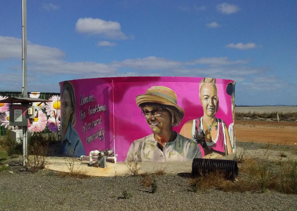 A vibrant magenta-pink corrugated metal structure adorned with large-scale portrait murals of two people, situated in an arid outback landscape. The building features striking street art with bold colors and appears to be a roadside attraction or community art installation in a remote desert region.
