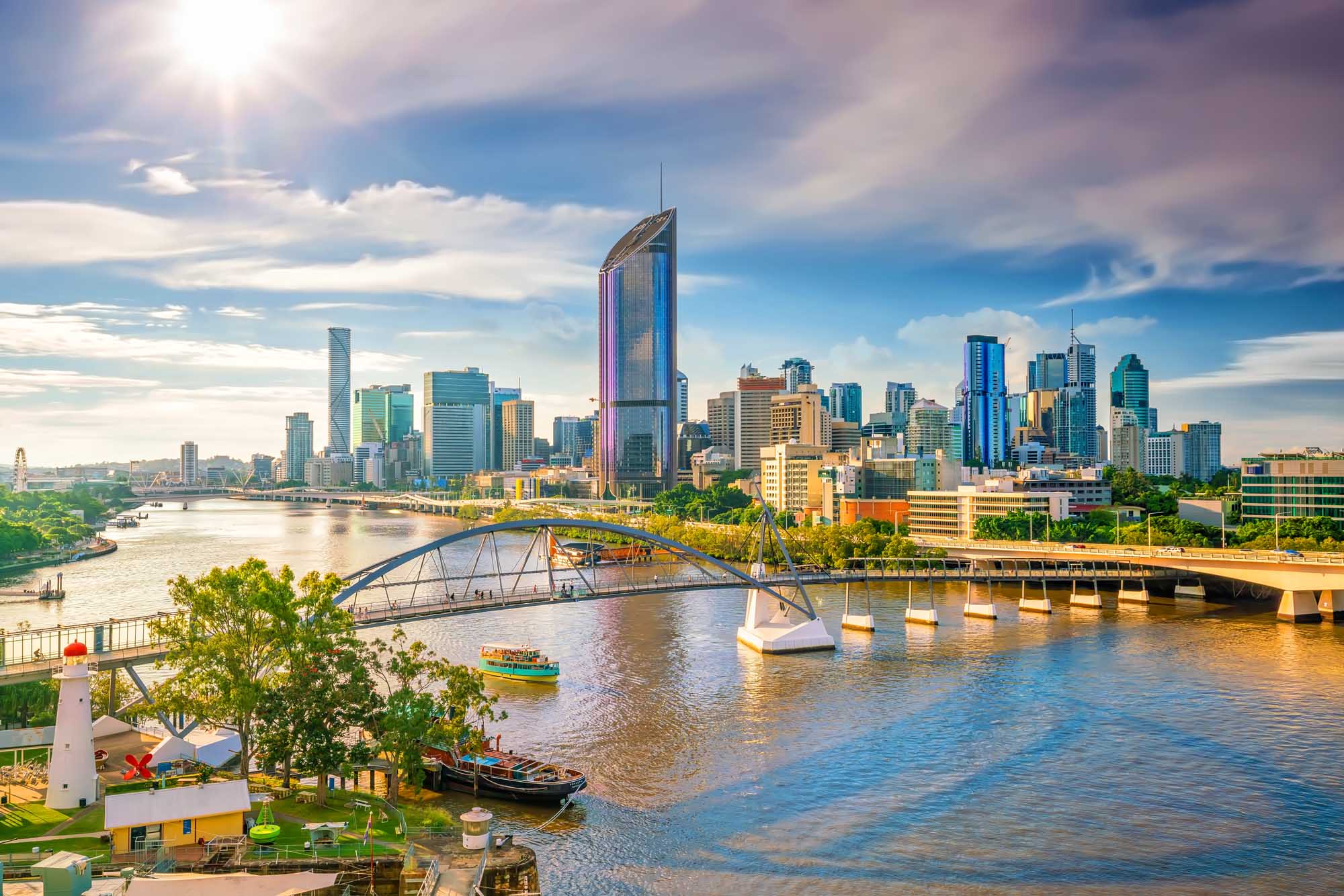 A vibrant aerial view of Brisbane's modern cityscape featuring the distinctive Story Bridge spanning the Brisbane River, with contemporary high-rise buildings bathed in golden sunset light, lush riverside parks, and small boats dotting the waterway under a partly cloudy sky.