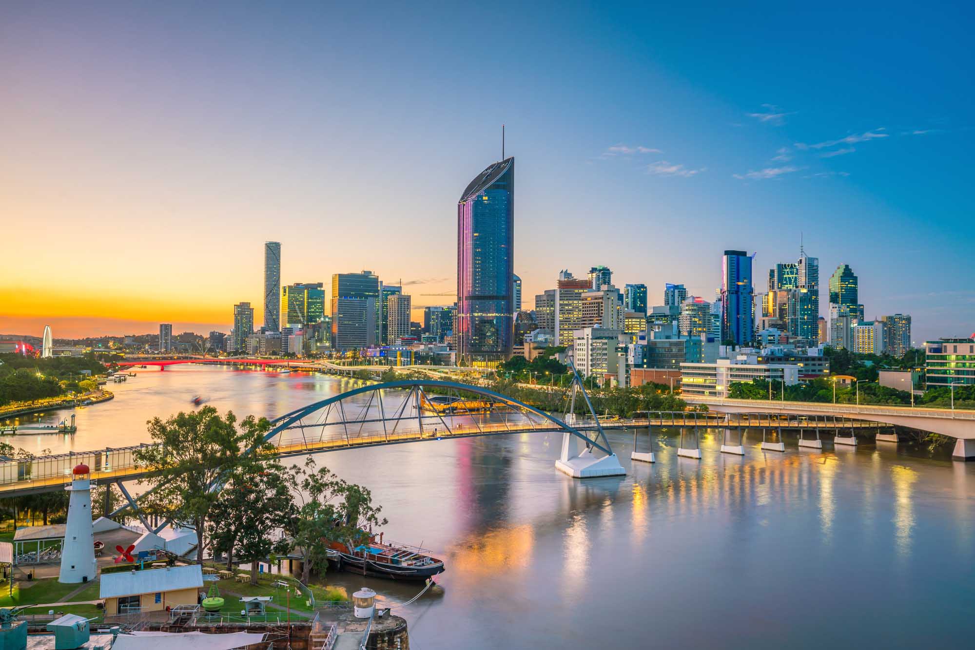 A stunning twilight view of Brisbane's modern skyline reflecting in the Brisbane River, featuring iconic skyscrapers, the distinctive Kurilpa Bridge in the foreground, and a riverside park with lighthouse. The golden and blue hour lighting creates a vibrant atmosphere across the cityscape and water.