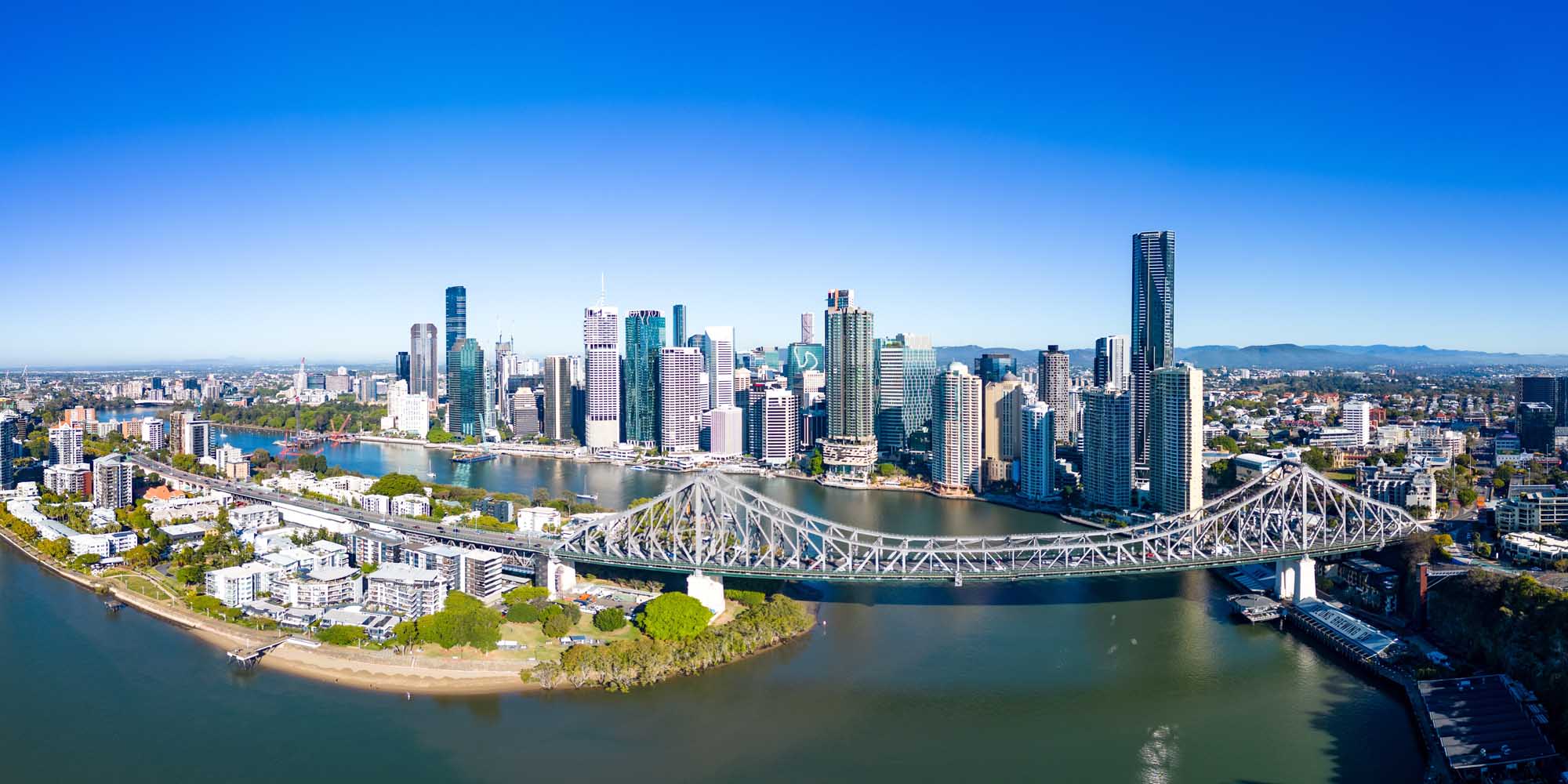 An aerial photograph of Brisbane's downtown skyline featuring the iconic Story Bridge spanning the Brisbane River, with the city's modern skyscrapers reflected in the water. The image captures the urban landscape from New Farm and Wilson Outlook Reserve on a clear, sunny winter morning in Queensland, Australia.