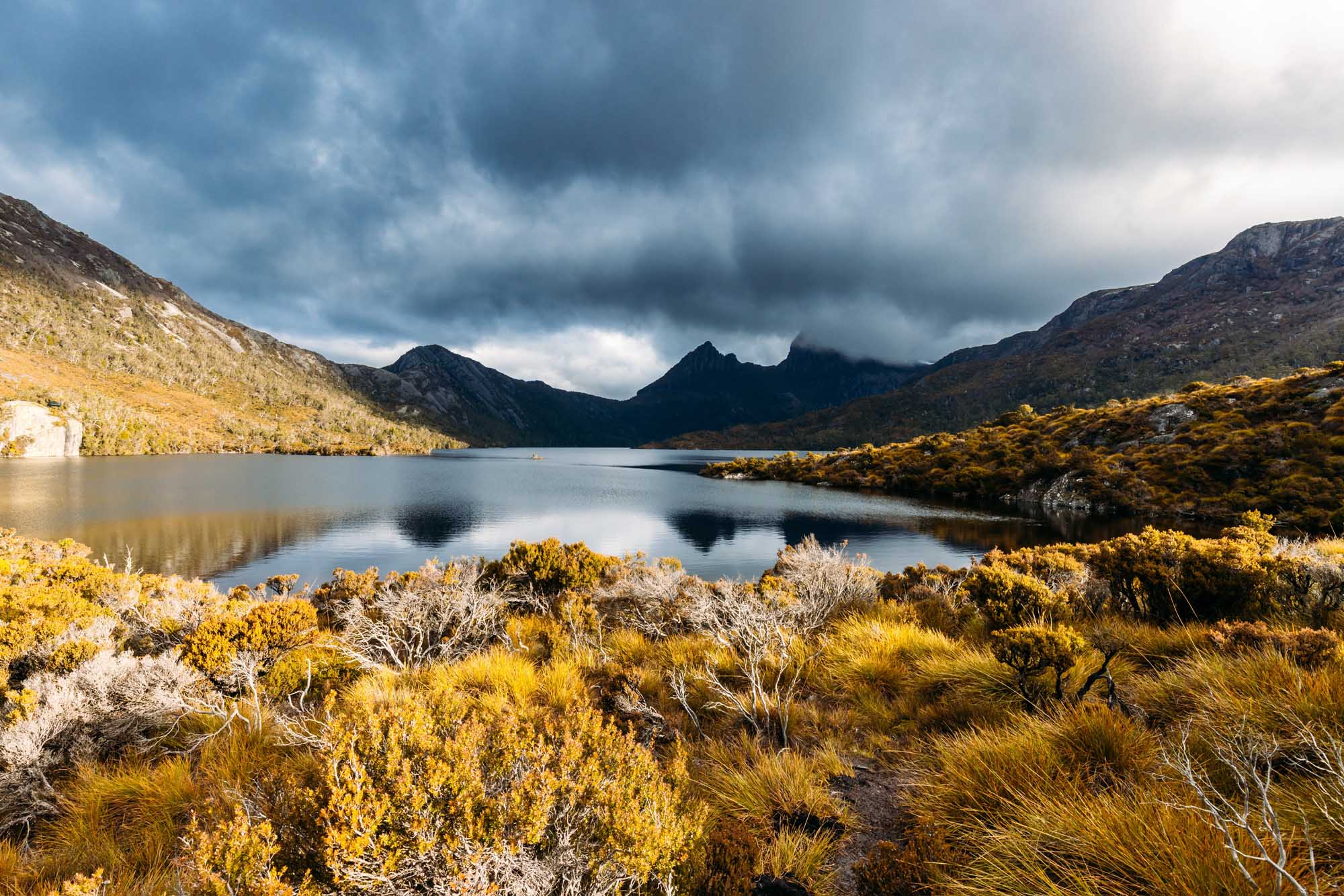 A stunning landscape view of Dove Lake with the iconic Cradle Mountain peaks reflected in the calm waters, surrounded by golden alpine vegetation and dramatic storm clouds gathering overhead during a cool spring afternoon near sunset.