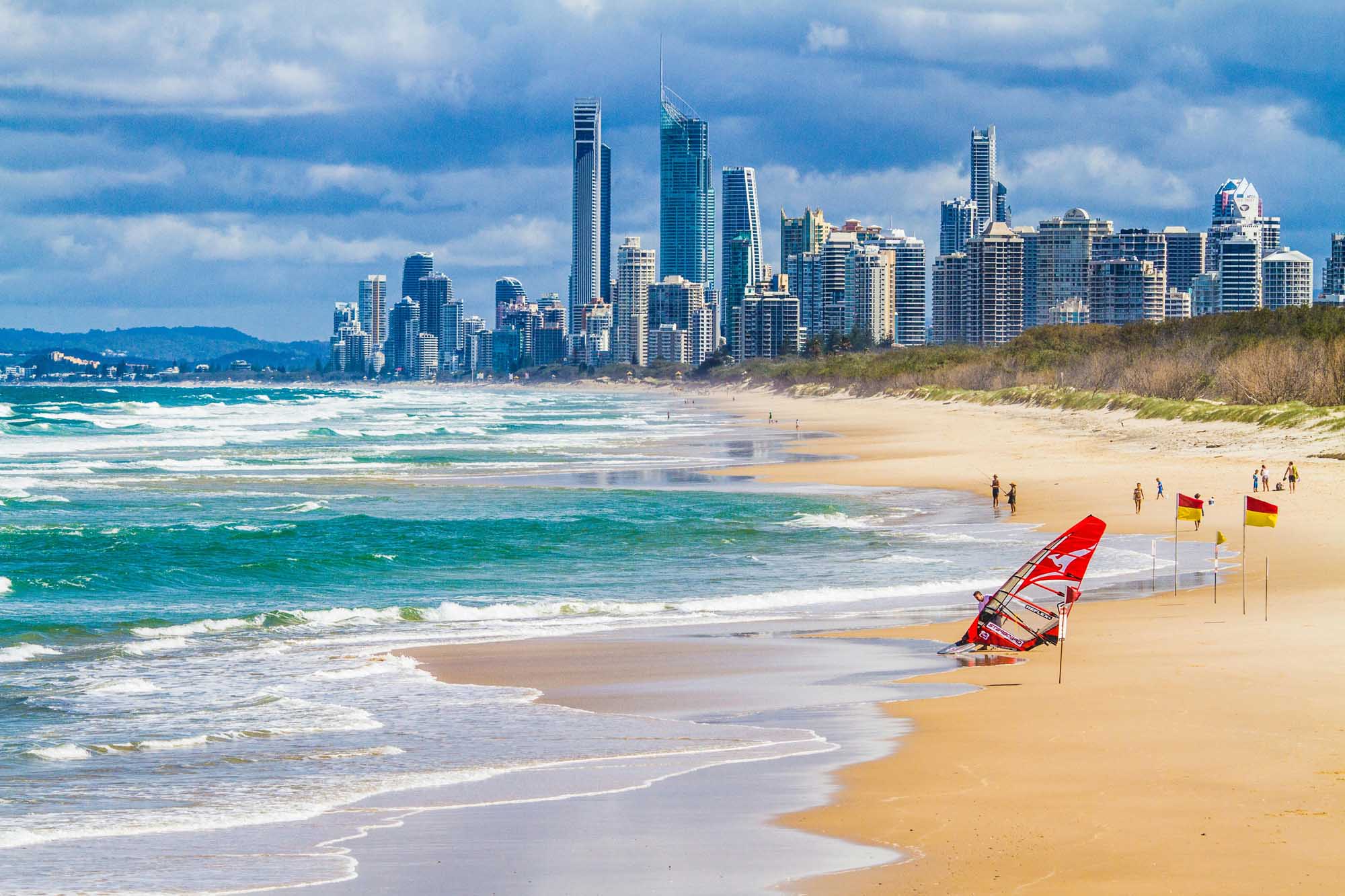 A vibrant coastal scene featuring a pristine sandy beach with turquoise waters and gentle waves in the foreground, a red windsurfing sail on the sand with lifeguard flags, scattered beachgoers, and a dramatic modern city skyline of high-rise buildings rising behind the dunes under a partly cloudy blue sky.