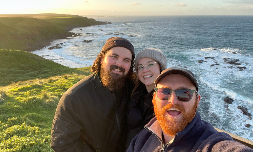 Three smiling visitors pose together at a dramatic coastal overlook featuring steep green cliffs, rocky shores, and crashing waves during golden hour. The scenic location showcases the rugged beauty of Australia's southern coastline with lush grass-covered headlands meeting the ocean below.