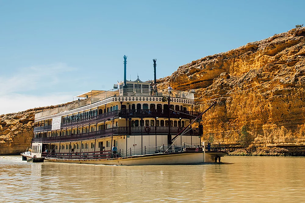 A classic multi-deck riverboat styled paddle-wheeler cruising along a scenic waterway, framed against towering golden limestone cliffs under clear skies. The vessel features traditional white and cream-colored superstructure with ornate railings and dual smokestacks, characteristic of inland river cruise ships.