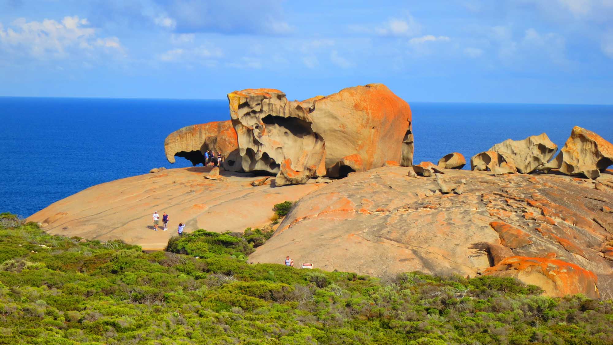 A stunning coastal landscape featuring the iconic Remarkable Rocks formation on Kangaroo Island, Australia. Large weathered granite boulders frame a pristine turquoise ocean under a clear blue sky, with visitors exploring the dramatic rocky outcropping and surrounding coastal terrain.