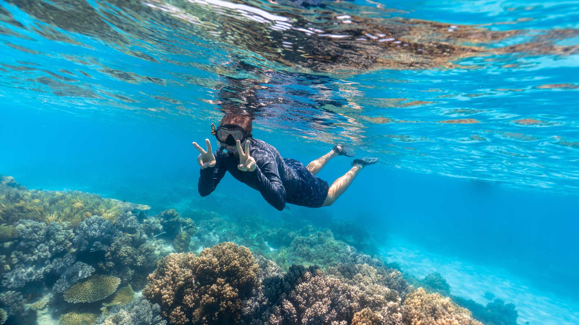 A young snorkeler in a wetsuit makes a peace sign while freediving above vibrant coral formations in the crystal-clear waters of the Great Barrier Reef. The underwater scene showcases diverse coral species and excellent visibility, capturing the beauty and accessibility of this iconic marine destination.