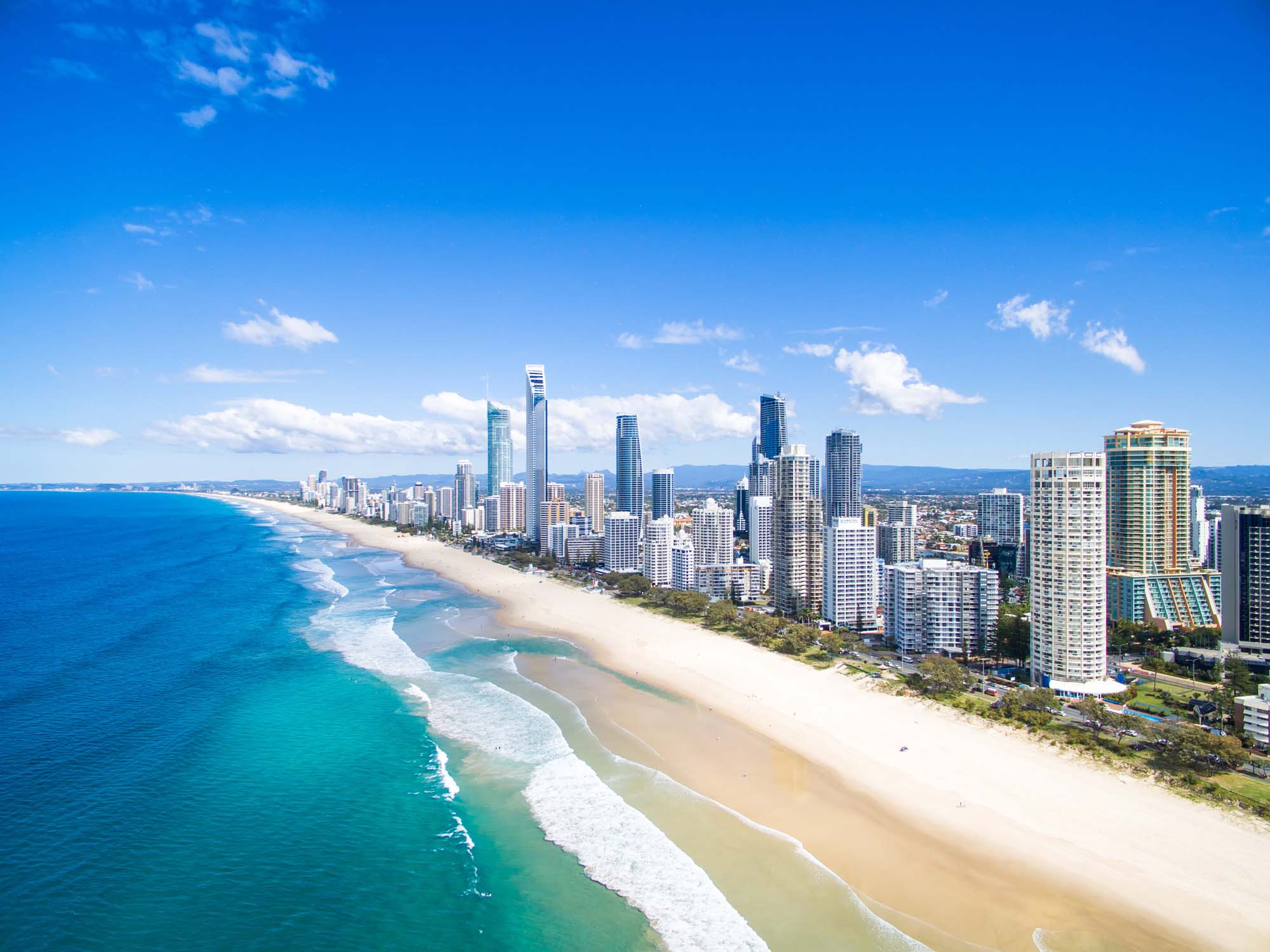 An aerial photograph of Surfers Paradise on Australia's Gold Coast, showing a pristine sandy beach stretching along a modern high-rise skyline with turquoise ocean waters and white-capped waves. The image captures the contrast between the natural coastal environment and the dense urban development characteristic of this popular vacation destination.