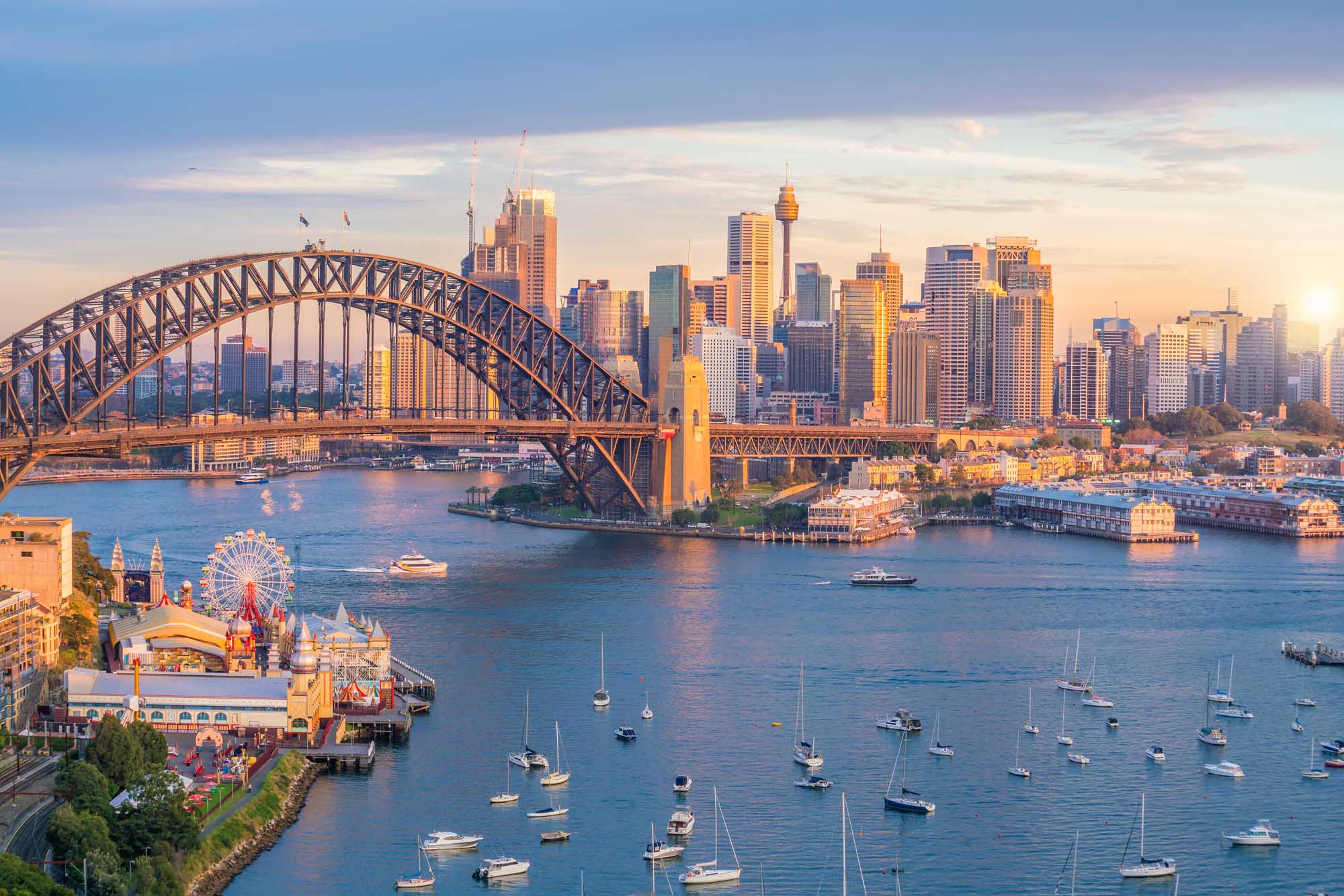 A stunning aerial view of Sydney's downtown skyline at golden hour, featuring the iconic Sydney Harbour Bridge spanning the blue waters, with sailboats dotting the harbour and Luna Park visible on the left foreground. The modern cityscape glows in warm light against a soft twilight sky, showcasing Australia's most recognizable landmarks.