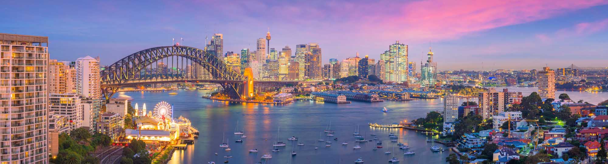 A breathtaking twilight panorama of Sydney's iconic skyline featuring the Sydney Harbour Bridge spanning across the glittering harbour, with the illuminated Opera House visible on the left, high-rise buildings reflecting golden light, and numerous sailboats dotting the calm waters during the magical blue hour.