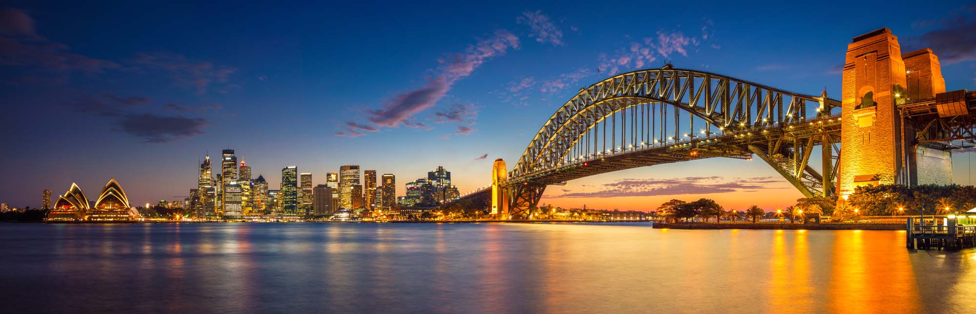 A stunning panoramic view of Sydney, Australia during the blue hour twilight, featuring the iconic Sydney Harbour Bridge illuminated in golden light, the distinctive Sydney Opera House on the left, and the glittering skyline reflected in the calm harbour waters.