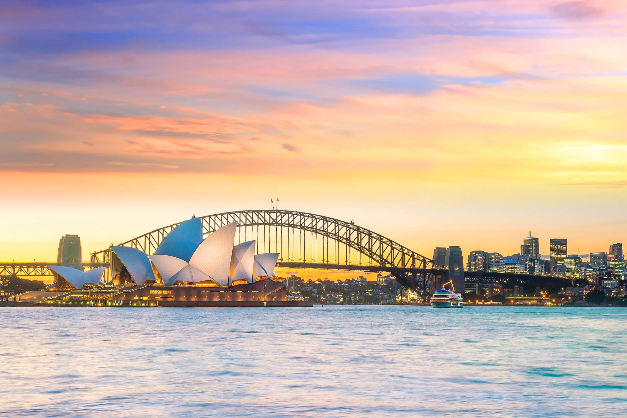 An iconic view of Sydney's most famous landmarks illuminated during golden hour, featuring the distinctive white shell-shaped Sydney Opera House in the foreground, the steel arch of the Sydney Harbour Bridge, and the city skyline glowing against a vibrant sunset sky reflected in the calm harbor waters.