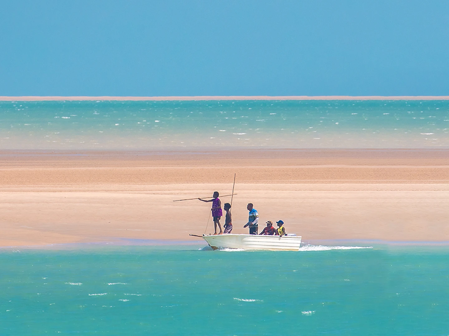 A small white fishing boat with several people aboard is beached in the shallow turquoise waters of Arnhem Land, with a pristine sandy shore and clear blue sky creating a serene tropical coastal scene.