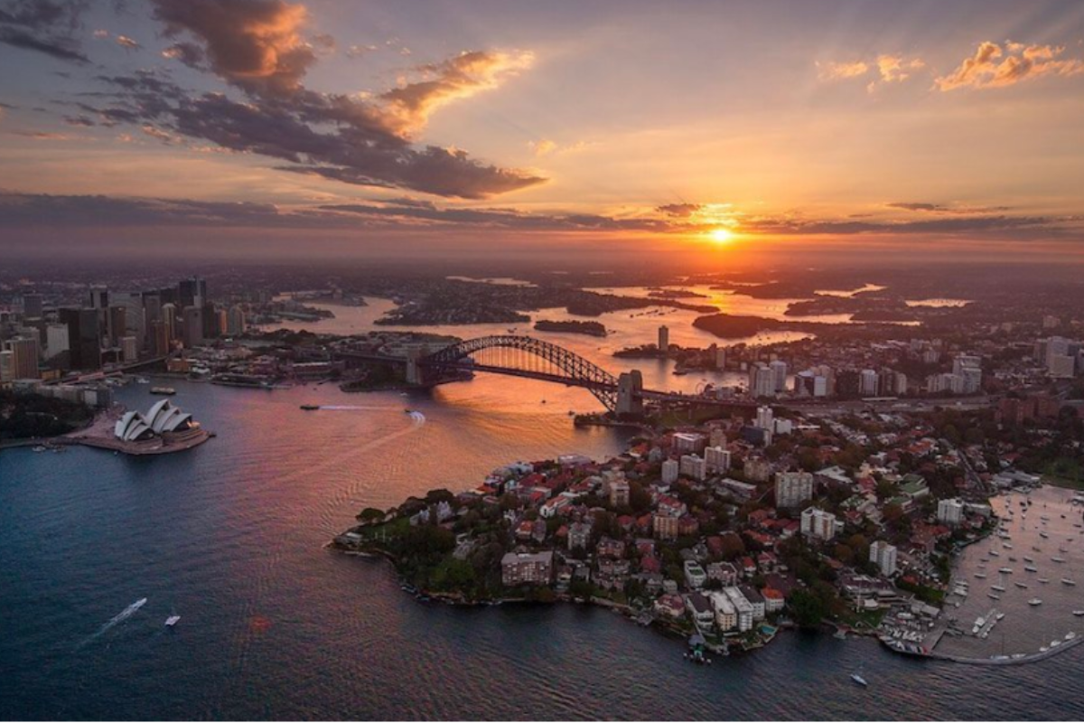 An aerial photograph of Sydney Harbour at sunset, capturing the iconic Sydney Opera House, Harbour Bridge, and surrounding cityscape bathed in golden light. The image showcases the city's distinctive landmarks and natural beauty across the water.