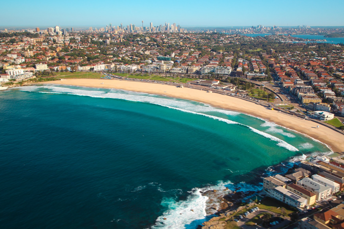 An aerial photograph of Bondi Beach in Sydney, Australia, showing the iconic golden sandy beach with turquoise waters, white-capped waves, and the surrounding beachside suburb with residential buildings, shops, and the Sydney cityscape with skyscrapers visible in the distance across the harbor.