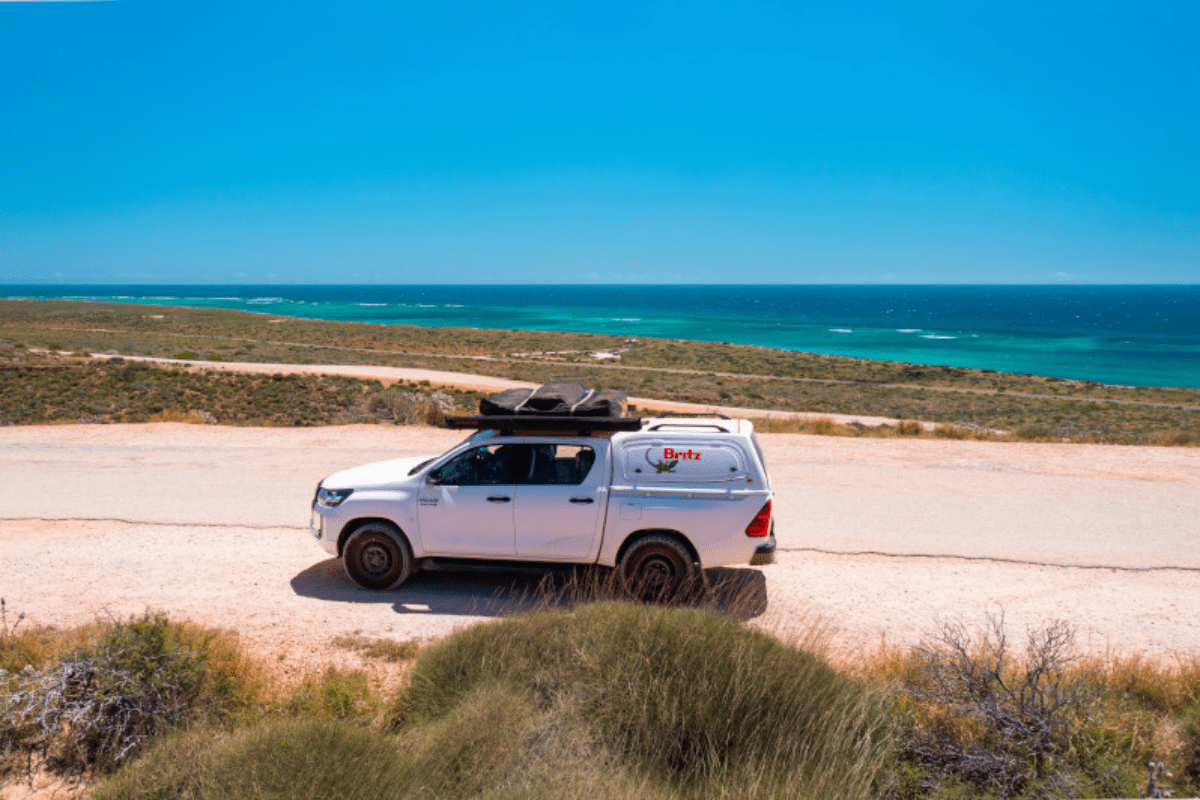 A white pickup truck equipped with a roof rack and storage canopy is parked on a sandy overlook with a panoramic view of turquoise ocean waters, coastal vegetation, and pristine beach below. The scene captures an adventurous camping or off-road travel experience in a remote coastal destination with clear blue skies.