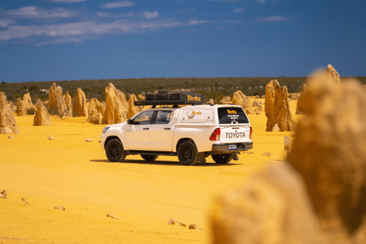 A white Toyota HiLux utility vehicle branded with Britz tour operator livery is parked among distinctive yellow limestone formations in Western Australia's Pinnacles Desert. The stark yellow sand and towering rock pillars create a dramatic desert landscape under clear blue skies.