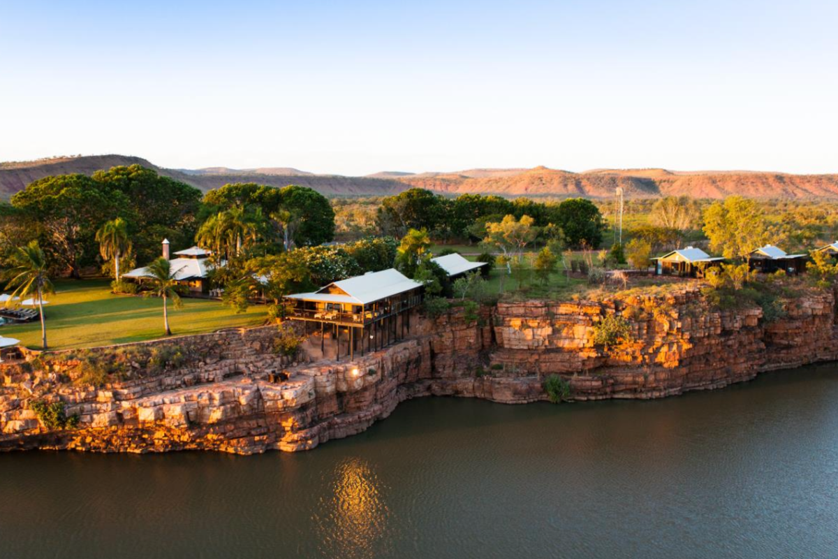 An exclusive riverside lodge perched on red sandstone cliffs overlooking a pristine waterhole, surrounded by native Australian vegetation including palm trees and eucalyptus, with distinctive white-roofed buildings and manicured lawns in a remote outback setting during golden hour.
