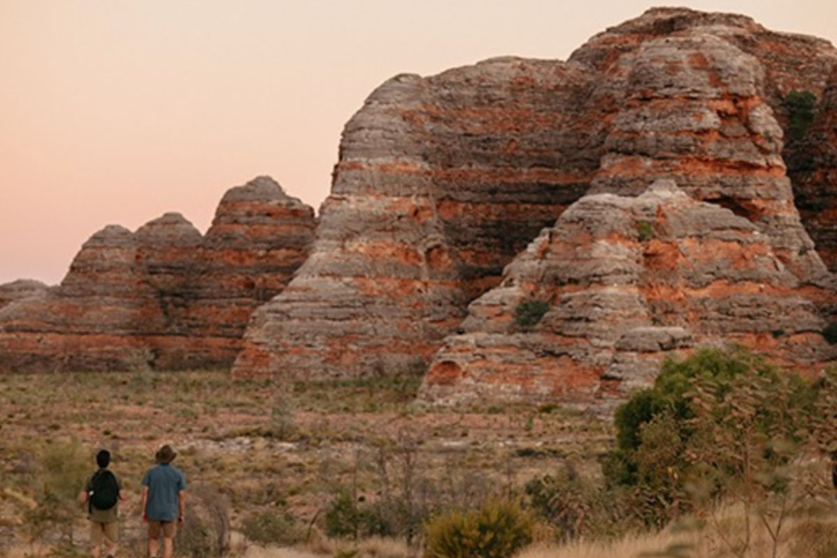 Two hikers stand in the foreground gazing at the distinctive striated rock formations of Bungle Bungle National Park, featuring dramatic layered sandstone domes with bands of red, orange, and grey. The arid landscape is dotted with sparse vegetation under a hazy, dusty sky characteristic of Australia's remote Kimberley region.