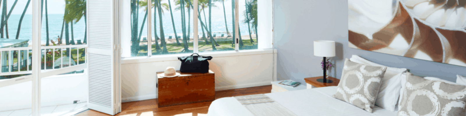 A hotel bedroom with a bed and wooden trunk, opening through folding doors onto a balcony overlooking a palm-lined beach