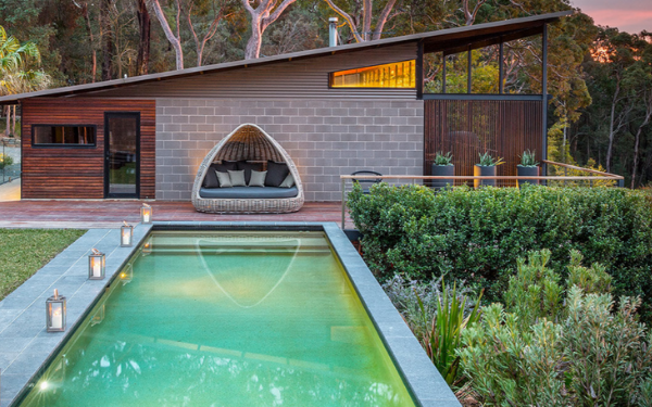 A contemporary residential retreat featuring a striking modern poolhouse with mixed material cladding (corrugated metal and gray brick), an integrated curved seating pod, and a rectangular illuminated pool with turquoise water. The property is nestled among eucalyptus trees with manicured hedging, creating a luxurious private sanctuary.