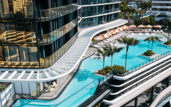 An aerial view of a contemporary high-rise residential building complex featuring multiple swimming pools with crystal-clear turquoise water, surrounded by modern glass and steel architecture, palm trees, and sunbathing areas with orange loungers.