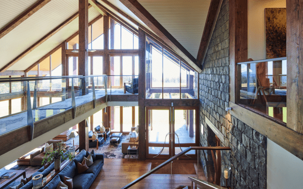 A stunning contemporary interior featuring a spacious double-height loft space with exposed wooden beams, floor-to-ceiling windows offering panoramic views, and a modern staircase connecting multiple levels. The space combines industrial architectural elements with luxury residential furnishings, creating an open-concept living environment.