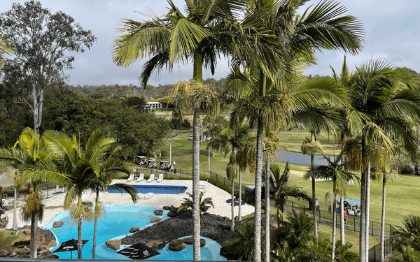 An expansive resort complex featuring a striking bright blue swimming pool with tropical landscaping, towering palm trees, and manicured green spaces overlooking a golf course and water body in the background, suggesting a premium leisure destination.