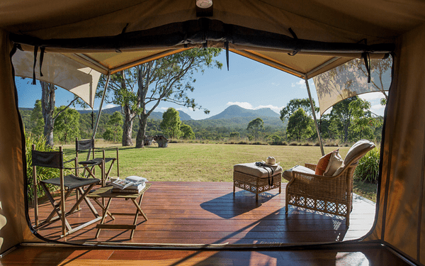 A view from inside a safari lodge tent showcasing a wooden deck with comfortable wicker seating, overlooking a vast grassy savanna with mountains in the distant background. The scene captures the essence of luxury safari accommodation with a blend of comfort and immersion in the natural landscape.