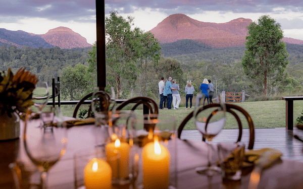 An elevated terrace view from a luxury resort or lodge featuring candlelit dining in the foreground with guests gathered on the lawn, overlooking a lush valley with distinctive red volcanic mountains in the distance under a dramatic cloudy sky.