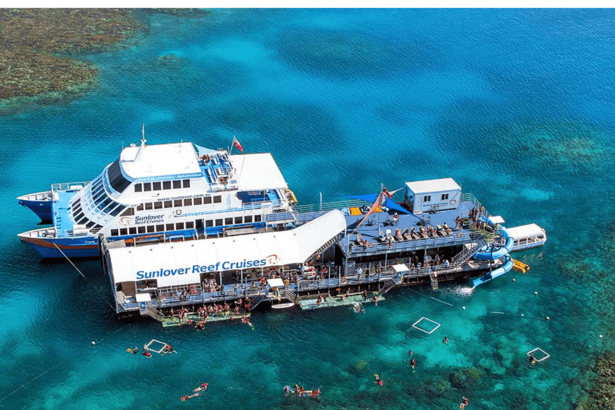 An aerial view of the Sunlover, a white and blue cruise vessel operated by Sunlover Reef Cruises, moored at a pontoon platform in the turquoise waters of the Great Barrier Reef. Multiple passengers are swimming and snorkeling in the crystal-clear water around the vessel, with visible underwater viewing platforms.
