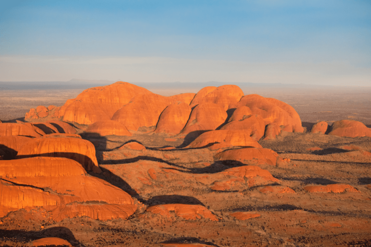 The iconic red rock formation of Uluru glows brilliant orange in the golden hour light, with deep shadows accentuating its layered geological structure. The vast arid landscape stretches to the horizon beneath a clear sky, capturing the dramatic natural beauty of Australia's most recognizable landmark.