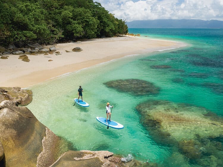Two paddleboarders enjoy crystal-clear turquoise waters off a pristine tropical beach, with lush green mangrove forest lining the shore and rocky outcrops visible in the shallow lagoon. The scene captures the natural beauty and water sports activities available in this idyllic destination.