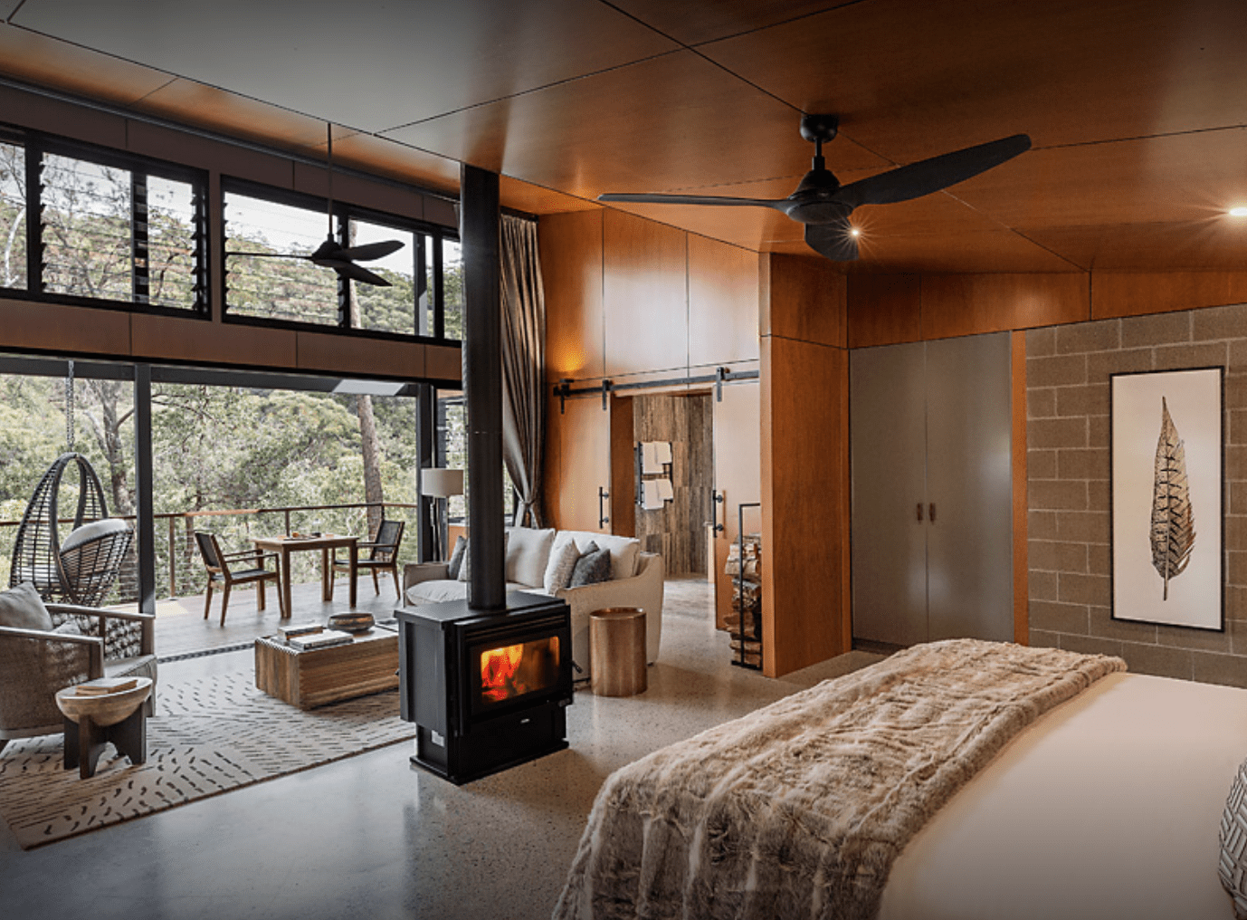 A sophisticated contemporary bedroom suite featuring warm wood paneling, a fireplace, and expansive windows overlooking a forested landscape. The open-concept design includes a sitting area with comfortable seating, a ceiling fan, and direct access to an outdoor terrace, exemplifying high-end resort or lodge accommodation.