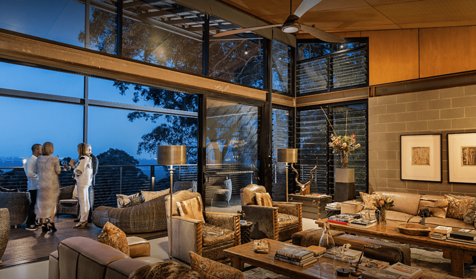 Guests socializing in a stylish lounge with high glass windows, leather furniture, and a night view over a treed hillside and distant water