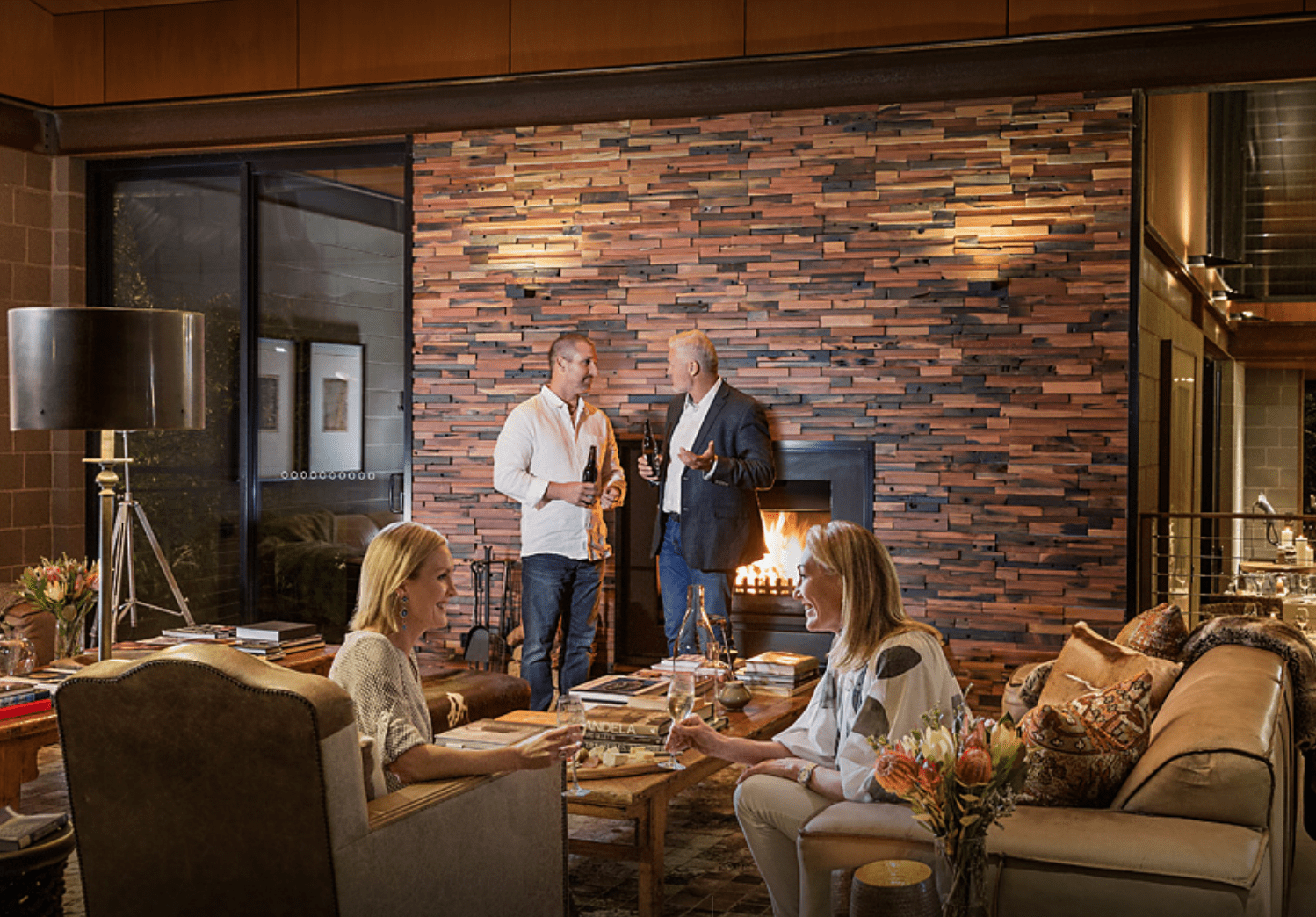 An elegant interior lounge space featuring a striking stacked brick fireplace accent wall with ambient lighting, where four adults socialize with drinks and books. The sophisticated room includes comfortable seating, a contemporary metal floor lamp, fresh floral arrangements, and modern architectural details.