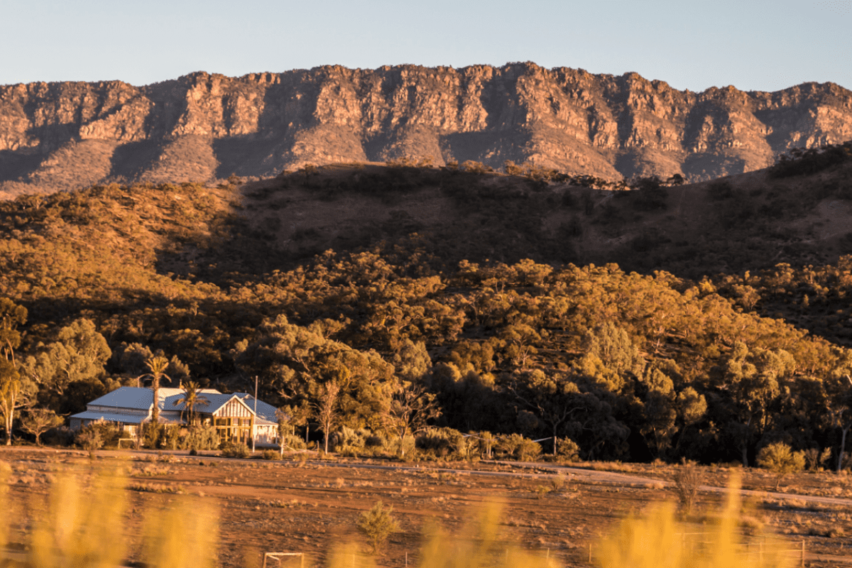 A picturesque rural homestead with a white house and palm tree nestled among golden eucalyptus trees along a waterway, set against a striking backdrop of dramatic cliff-faced mountains illuminated by golden hour sunlight.