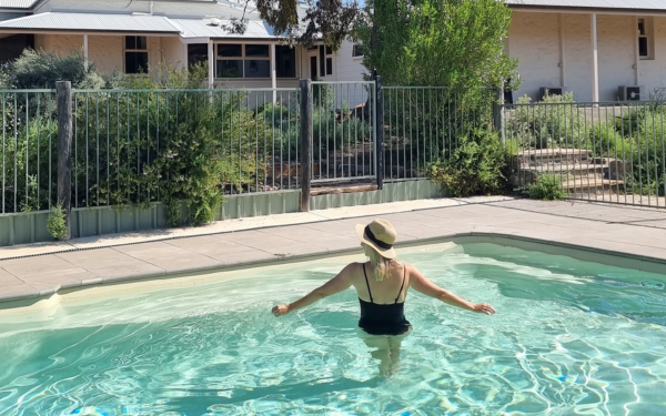A woman wearing a hat and swimsuit enjoys a sunny day in a clear turquoise swimming pool at a residential property. The backyard features metal safety fencing, mature landscaping with trees and shrubs, and a single-story house with a covered patio area in the background.