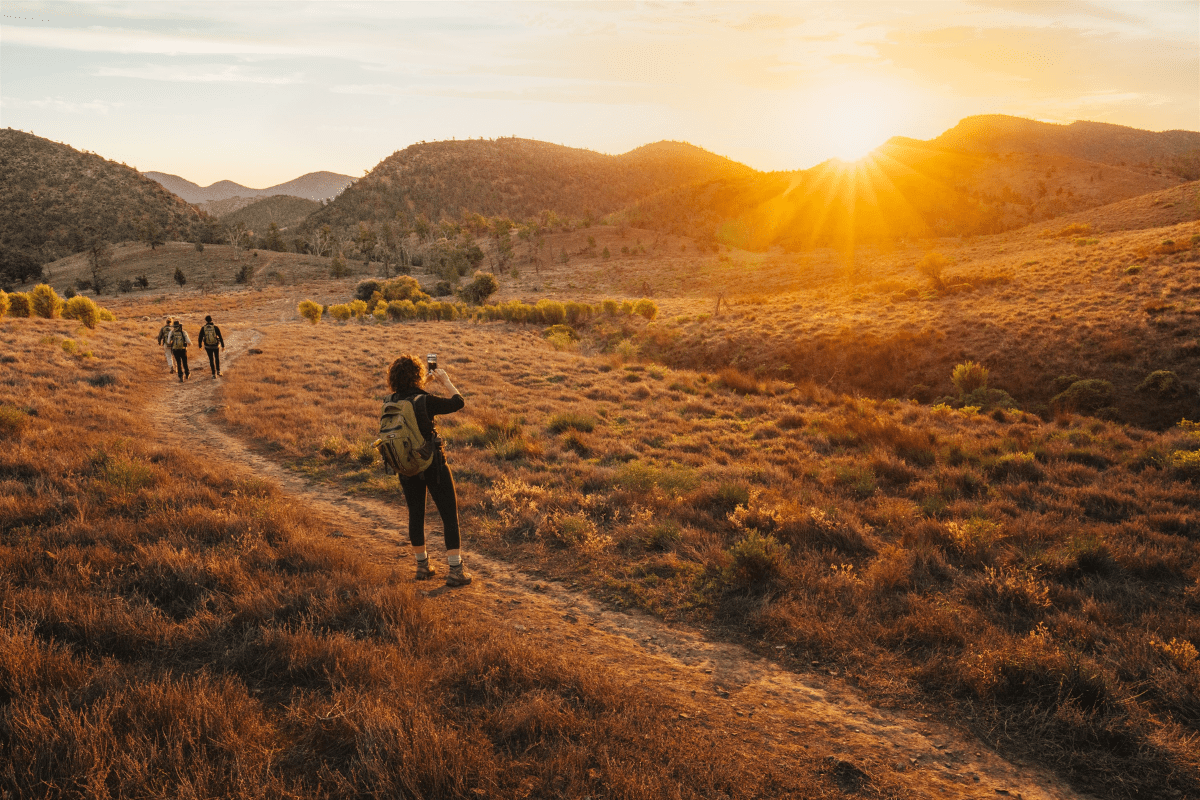 A group of hikers explores a vast, arid mountain valley during golden hour, with one hiker in the foreground capturing the scenic landscape while others trek along a trail. The warm, golden sunlight bathes the terrain, shrubland, and distant mountains in a spectacular sunset glow.