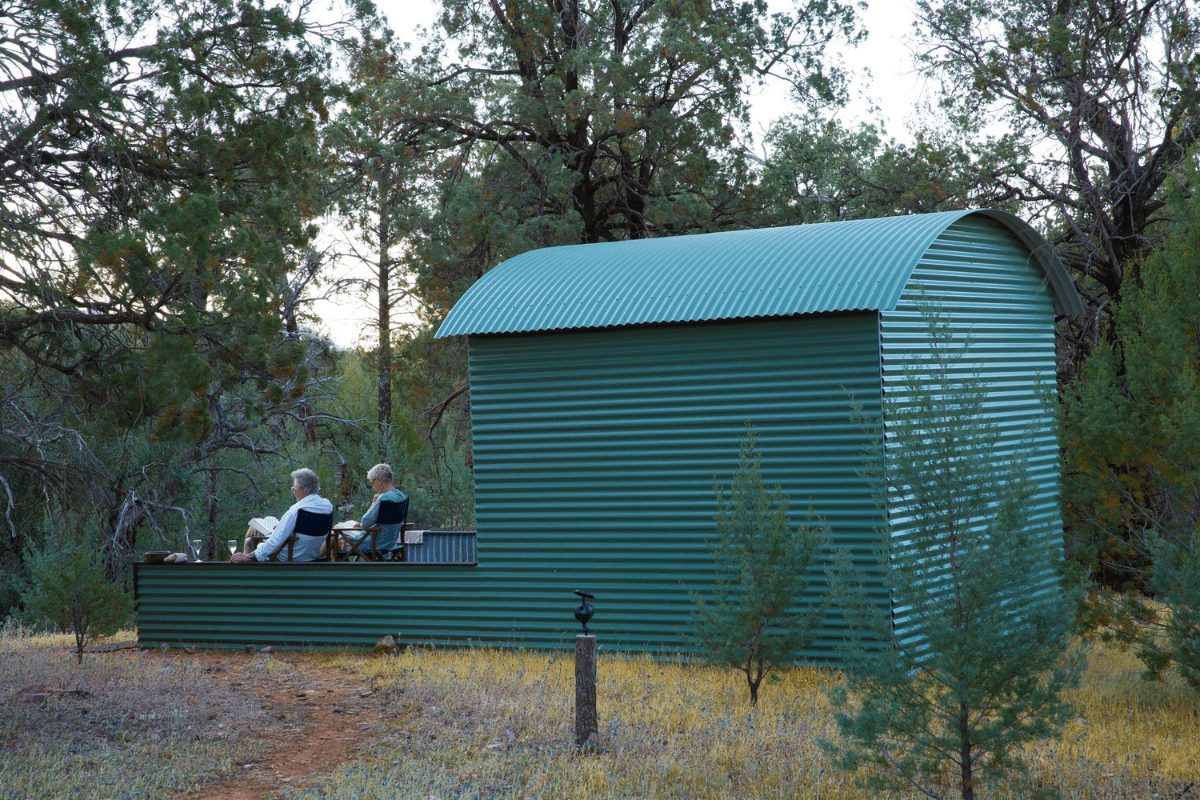 A distinctive turquoise corrugated metal structure with a curved barrel roof nestled among tall pine trees in a dry, golden grassland setting. Two people can be seen sitting on a front deck, appearing to enjoy the peaceful wilderness surroundings.
