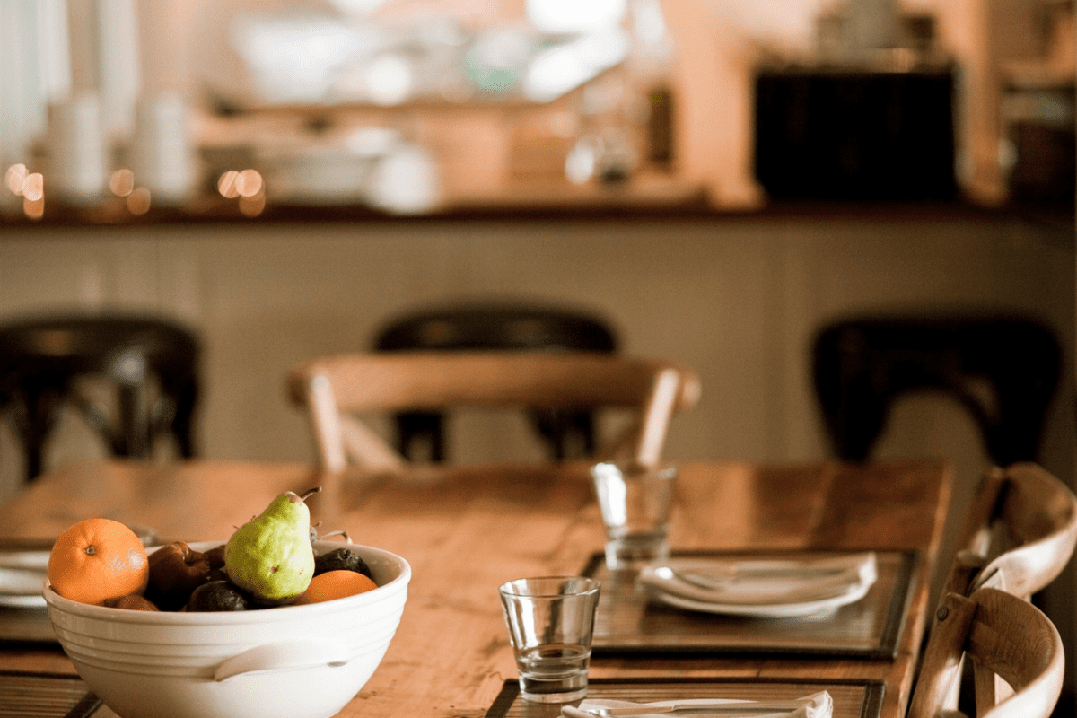A sophisticated dining space featuring a wooden table set with place settings, glasses of water, and white napkins. A prominent white ceramic bowl filled with fresh oranges, a pear, and dark fruits is prominently displayed in the foreground, while the background shows a blurred kitchen area with warm ambient lighting and neutral tones.