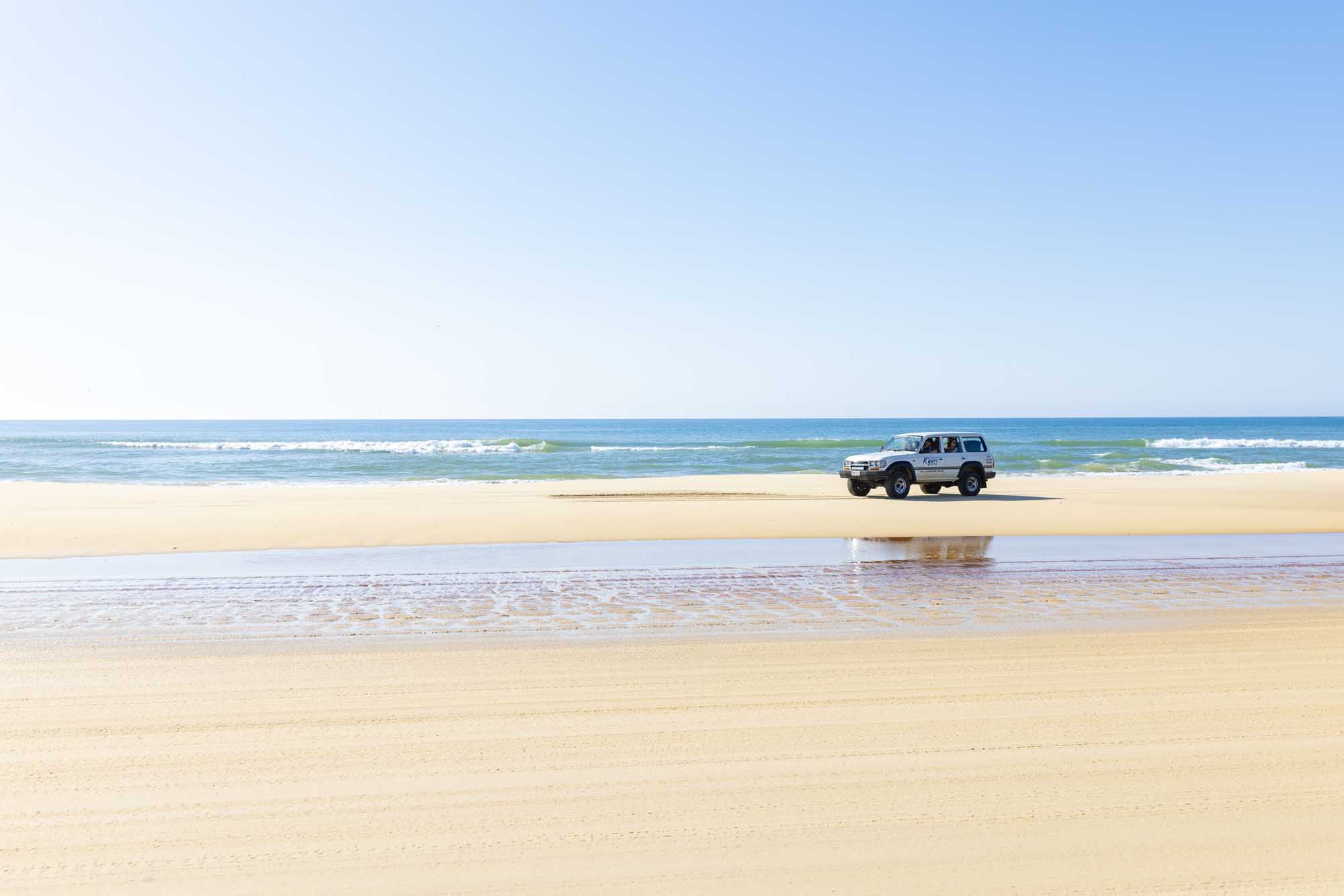 A white four-wheel drive vehicle travels along a pristine sandy beach on K'gari Fraser Island, Queensland, with turquoise ocean waters and gentle waves in the background under a clear blue sky. The image captures the essence of an off-road island adventure along Australia's famous coastal landscape.