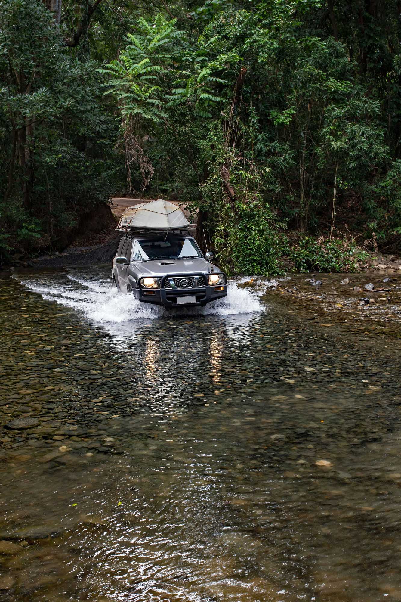 A rugged SUV equipped with a roof-mounted tent fords through a shallow river in a dense tropical rainforest setting, showcasing adventure travel and off-road exploration in an Australian wilderness environment.
