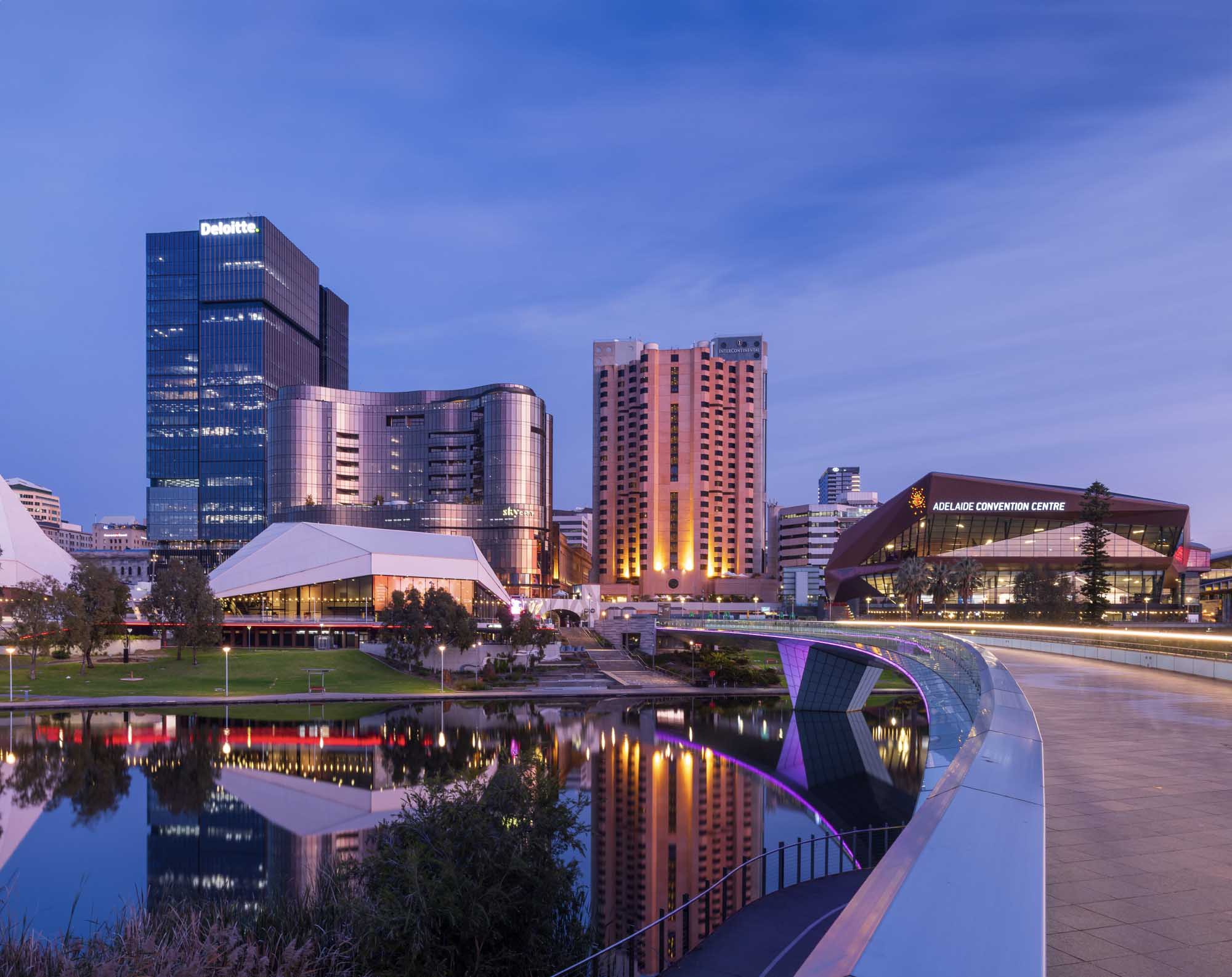 A stunning twilight view of Adelaide's modern waterfront precinct featuring the illuminated Adelaide Convention Centre, contemporary curved architecture, high-rise hotels, and the Deloitte office tower reflected in calm water. The scene captures the vibrant urban landscape of South Australia's capital city during the blue hour with architectural lighting highlighting the buildings' distinctive designs.