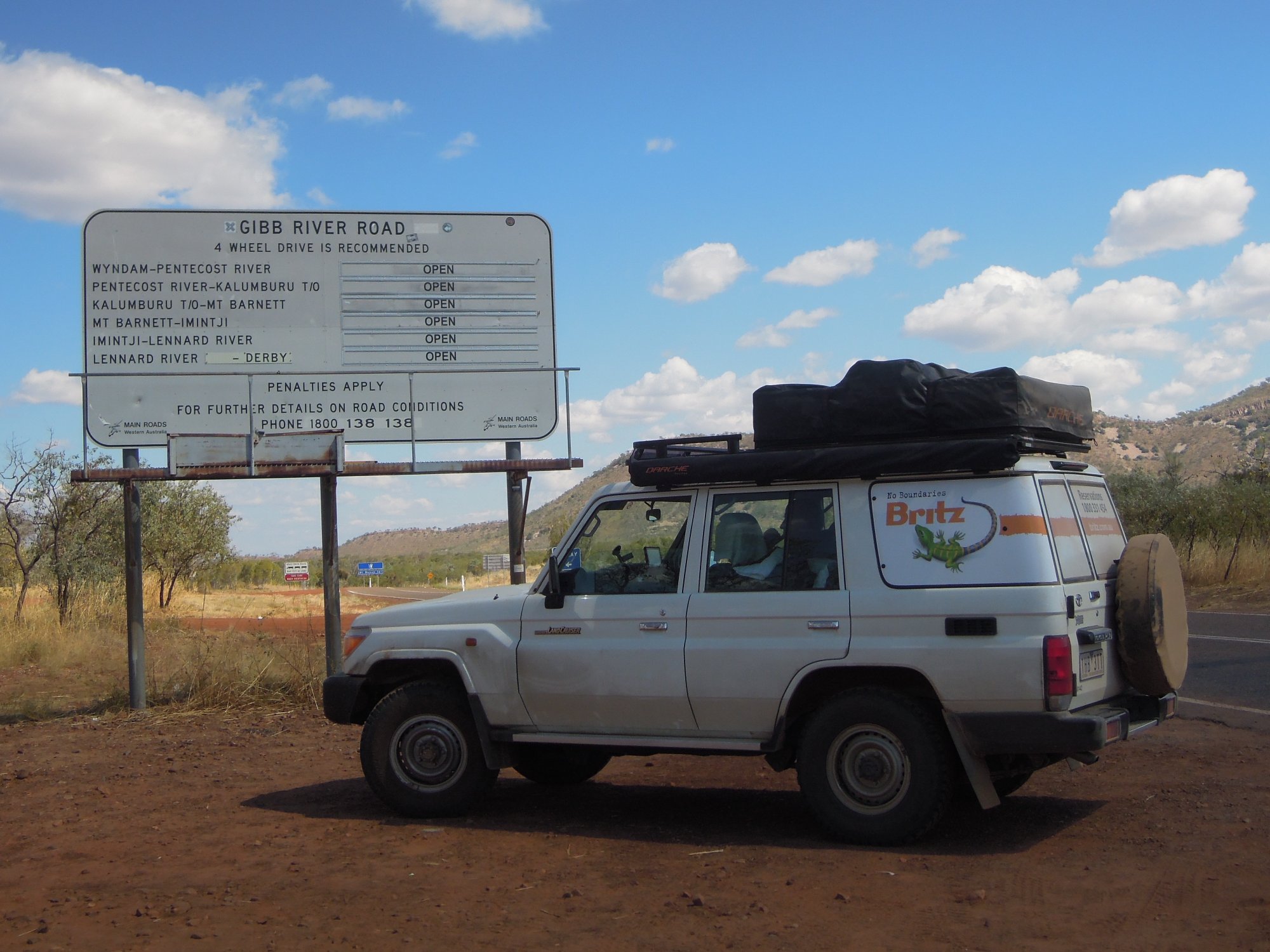 A white four-wheel-drive vehicle with roof rack and spare tire is parked at the beginning of Gibb River Road in Western Australia's remote Kimberley region. The scene shows the iconic road sign displaying various river crossing conditions and 4WD drive recommendations, with rugged bushland and rocky hills visible in the background under a clear blue sky.