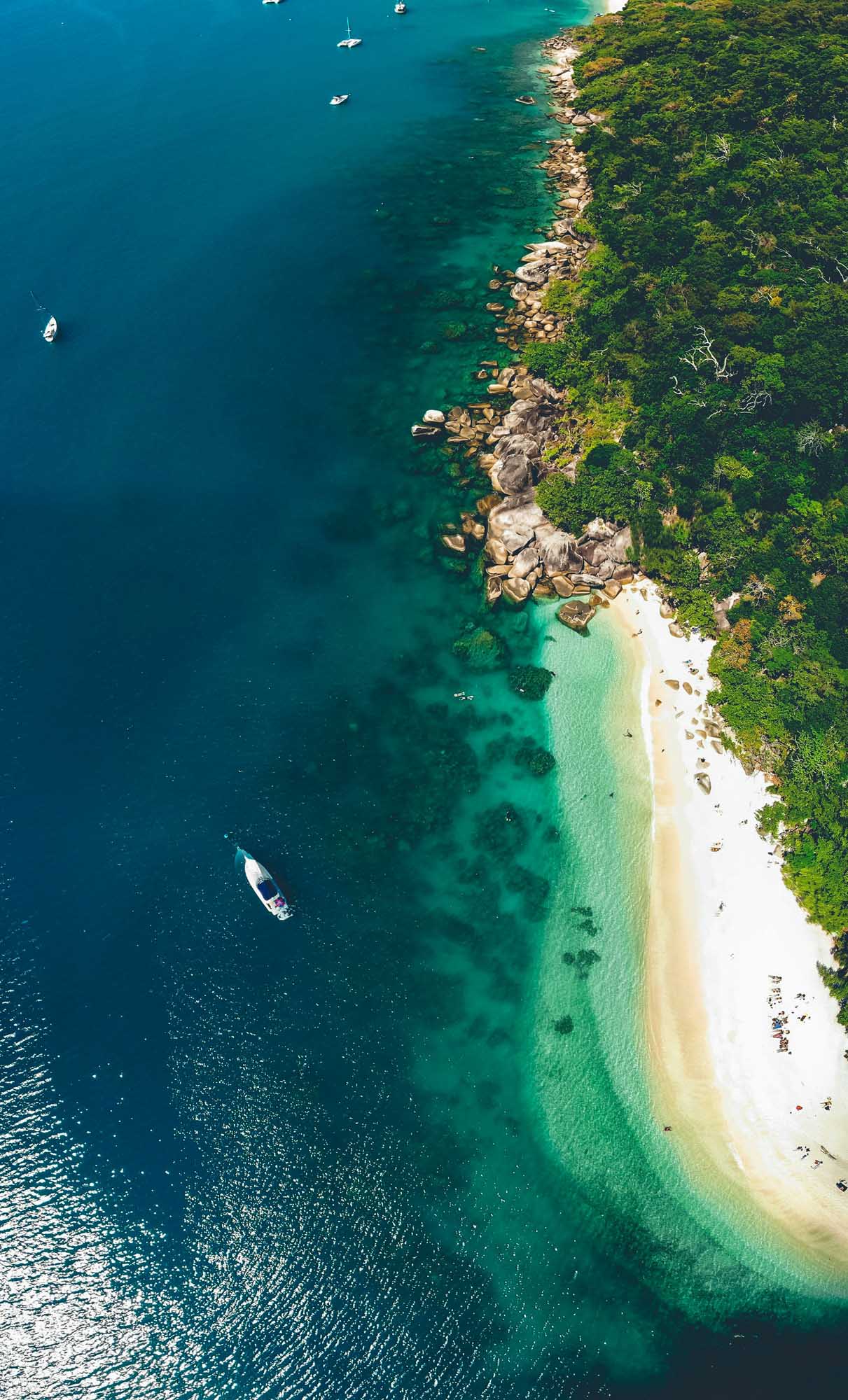 A breathtaking aerial photograph of Fitzroy Island showing pristine turquoise waters, a white sandy beach, lush tropical rainforest vegetation, and scattered anchored boats. The image captures the dramatic contrast between the deep blue ocean, shallow turquoise shallows, rocky granite outcrops, and dense green jungle coastline.