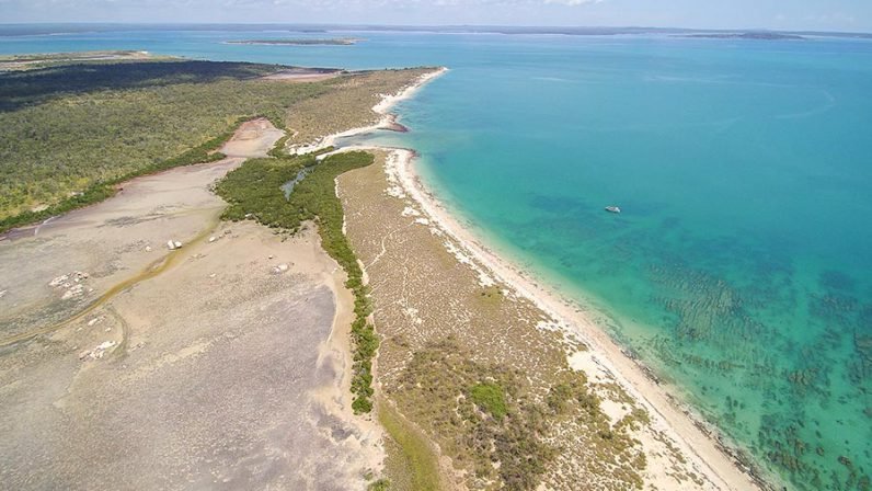 An aerial photograph of Australia's remote Kimberley coastline, featuring pristine turquoise waters, white sandy beaches, and dense tropical vegetation meeting rugged terrain. The image showcases the dramatic contrast between the arid inland landscape and the vibrant coastal waters, with small vessels visible offshore.