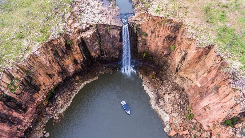 An aerial drone shot capturing a small expedition vessel navigating a pristine freshwater pool beneath a dramatic waterfall in the Kimberley region. The surrounding landscape features distinctive red rock formations and sparse vegetation, showcasing the remote natural beauty of this iconic Australian wilderness destination.