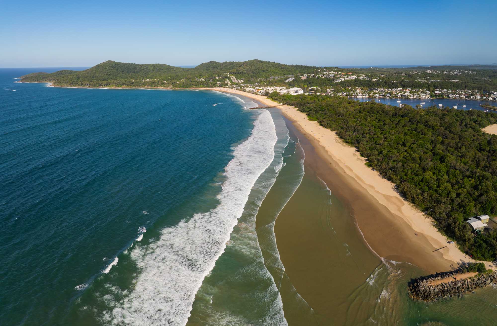 An aerial photograph of Noosa Heads on Australia's Sunshine Coast, Queensland, showcasing pristine sandy beaches, turquoise ocean waters with white-capped waves, a river entrance with distinctive water patterns, dense coastal vegetation, and residential development along the shoreline under clear blue skies.