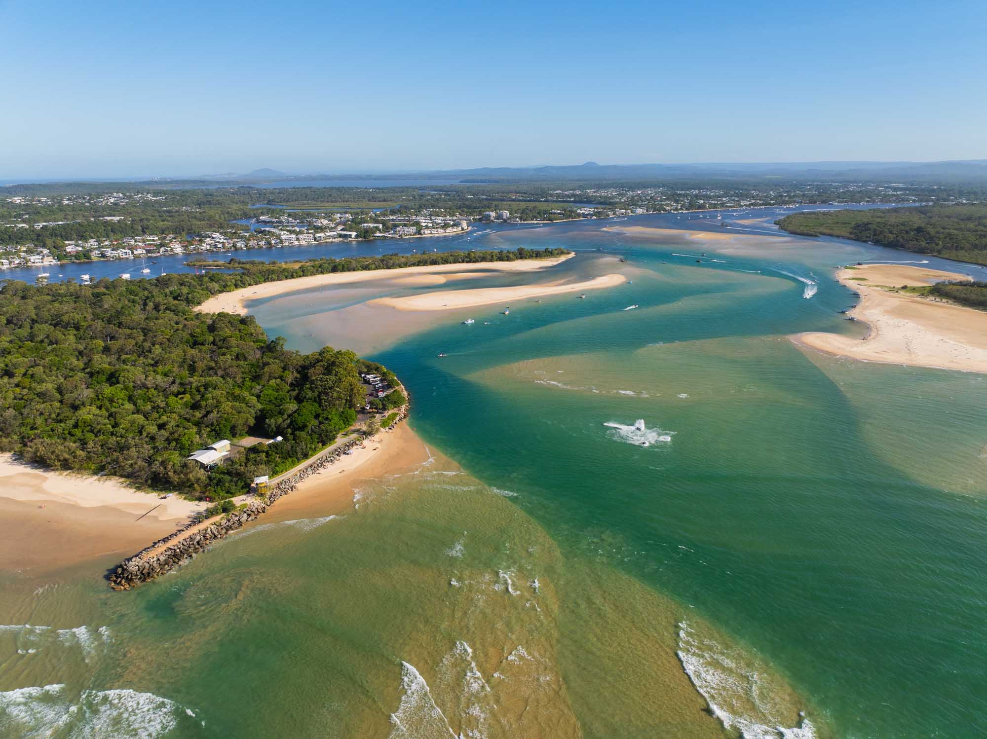 Stunning aerial photograph of Noosa Heads on Queensland's north shore, showcasing the pristine river entrance with turquoise waters, sandy sandbars, lush coastal vegetation, and waterfront residences. The image captures the natural beauty of this popular Australian coastal destination with its distinctive beach formations and waterways.