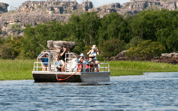 A group of tourists aboard a shallow-draft flat-bottomed boat navigates a scenic river landscape, with dramatic rocky outcrops and green vegetation visible in the background. The boat appears to be conducting an eco-tourism or nature expedition tour in a remote or protected natural area.